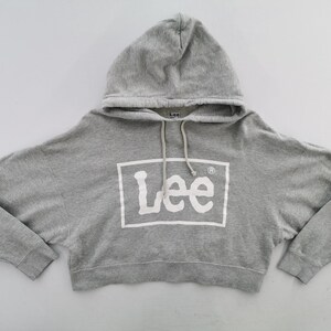 May include: A grey cropped hoodie with a drawstring hood. The front displays the white "Lee" logo within a white rectangle. The hoodie has long sleeves and a cropped waist. The fabric appears to be a soft, comfortable material.
