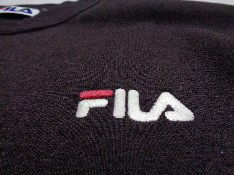 Pu&ograve; includere: Una felpa viola scuro con il logo del marchio FILA ricamato sul petto. Il logo &egrave; composto dalla parola FILA in filo bianco con una barra orizzontale rossa sopra la lettera I. Un'etichetta parziale del marchio &egrave; visibile sullo scollo.