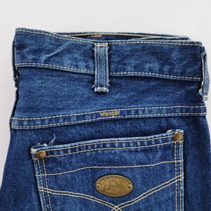 May include: Close-up of a pair of blue denim Wrangler jeans. The jeans feature a classic five-pocket design with a Wrangler label on the back pocket. The stitching is a contrasting light color, and the jeans have a vintage look.