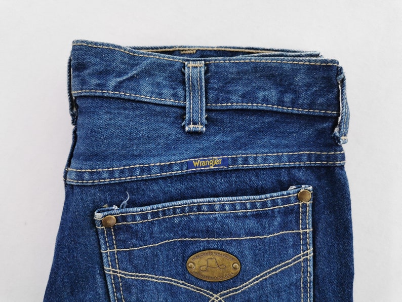May include: Close-up of a pair of blue denim Wrangler jeans. The jeans feature a classic five-pocket design with a Wrangler label on the back pocket. The stitching is a contrasting light color, and the jeans have a vintage look.