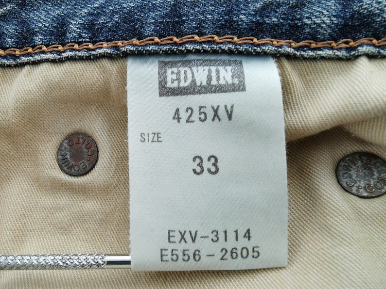 Puede incluir: Primer plano del interior de unos vaqueros azules. Una etiqueta blanca muestra "EDWIN", "425XV" y la talla "33". Se ven dos botones met&aacute;licos, uno con "EDWIN CO.". Los vaqueros son de estilo denim cl&aacute;sico.