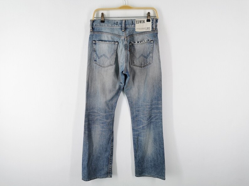 May include: Light blue denim jeans with a faded wash, hanging on a wooden hanger. The jeans have a classic five-pocket design, a straight leg, and the brand name "EDWIN" is visible on the back pocket.