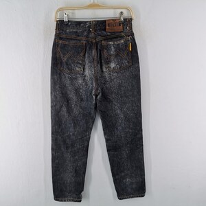 May include: Acid wash denim jeans with a classic five-pocket design. The jeans feature a faded black wash with orange stitching and a leather patch on the back. The jeans are hanging on a hanger against a white background.