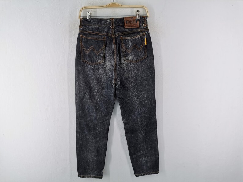 May include: Acid wash denim jeans with a classic five-pocket design. The jeans feature a faded black wash with orange stitching and a leather patch on the back. The jeans are hanging on a hanger against a white background.