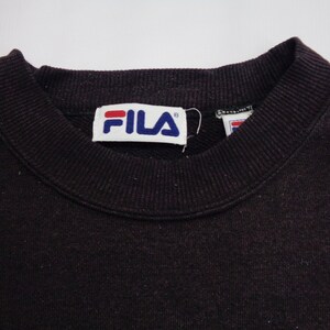 Pu&ograve; includere: Una felpa girocollo marrone scuro con colletto a coste e un'etichetta rettangolare bianca del marchio all'interno dello scollo. L'etichetta riporta la scritta FILA in lettere blu con una barra orizzontale rossa sopra la lettera F.
