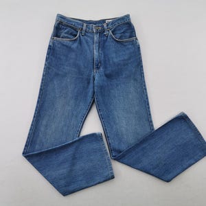 May include: Blue denim jeans with a classic five-pocket design. The jeans have a straight leg and a button and zipper closure. The jeans are made of a medium-wash denim fabric. The jeans are laid flat on a white surface.