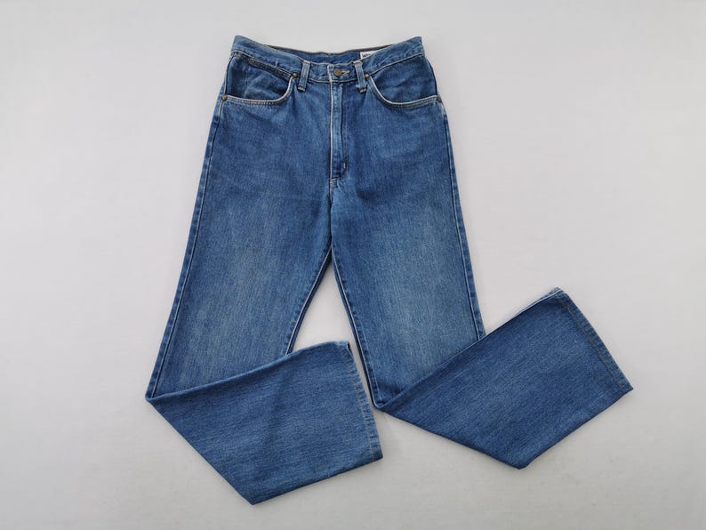 May include: Blue denim jeans with a classic five-pocket design. The jeans have a straight leg and a button and zipper closure. The jeans are made of a medium-wash denim fabric. The jeans are laid flat on a white surface.