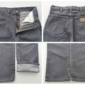 May include: Close-up shots of gray denim jeans. The jeans have a button and zipper closure, front and back pockets, and a leather patch. The bottom hem is cuffed, revealing the lighter fabric inside. The Wrangler brand is visible.