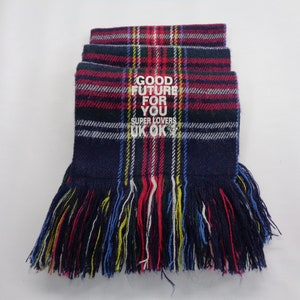 Puede incluir: Una bufanda doblada con estampado de cuadros en azul marino, rojo y verde con texto blanco que dice GOOD FUTURE FOR YOU SUPER LOVERS UK OK. La bufanda presenta un borde con flecos de hilos multicolores en rojo, azul, amarillo y blanco.