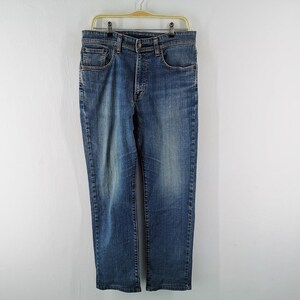 May include: Blue denim jeans with a classic five-pocket design, hanging on a gold hanger. The jeans have a straight leg cut and a button and zip closure. The denim has a faded wash effect.