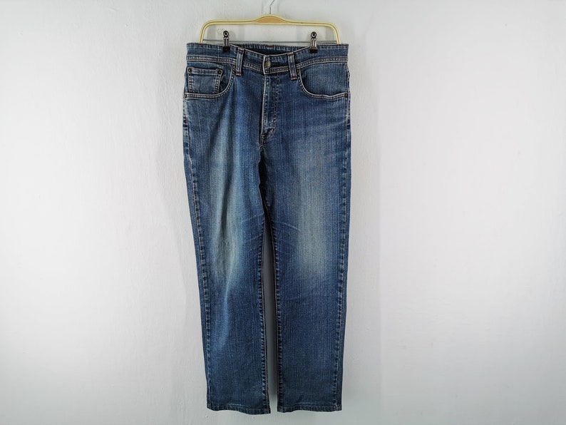 May include: Blue denim jeans with a classic five-pocket design, hanging on a gold hanger. The jeans have a straight leg cut and a button and zip closure. The denim has a faded wash effect.