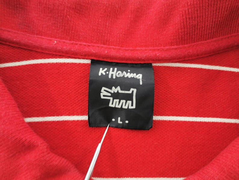 Puede incluir: Primer plano de un polo rojo con rayas horizontales blancas. La camisa presenta una etiqueta negra con el texto "K. Haring" y un gr&aacute;fico de perro estilizado. La etiqueta tambi&eacute;n indica una talla "L".