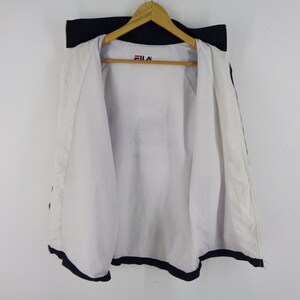 May include: White and navy blue Fila jacket hanging on a wooden hanger. The jacket is open, revealing a white interior. The Fila logo is visible on the upper chest area. The jacket has navy blue trim.