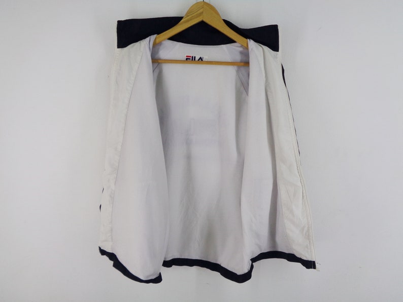 May include: White and navy blue Fila jacket hanging on a wooden hanger. The jacket is open, revealing a white interior. The Fila logo is visible on the upper chest area. The jacket has navy blue trim.