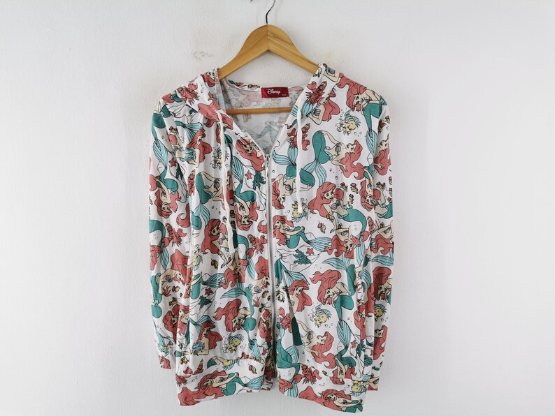 May include: White zip-up hoodie featuring a repeating pattern of Ariel from Disney's The Little Mermaid. The design includes Ariel, sea creatures, and seaweed in shades of red, teal, and white. The hoodie has a hood and a full-length zipper.
