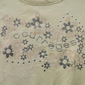 May include: A light yellow t-shirt with a floral design. The design includes gray and light pink flowers, with the word "courreges" in gray lettering. Small silver studs are scattered throughout the design.