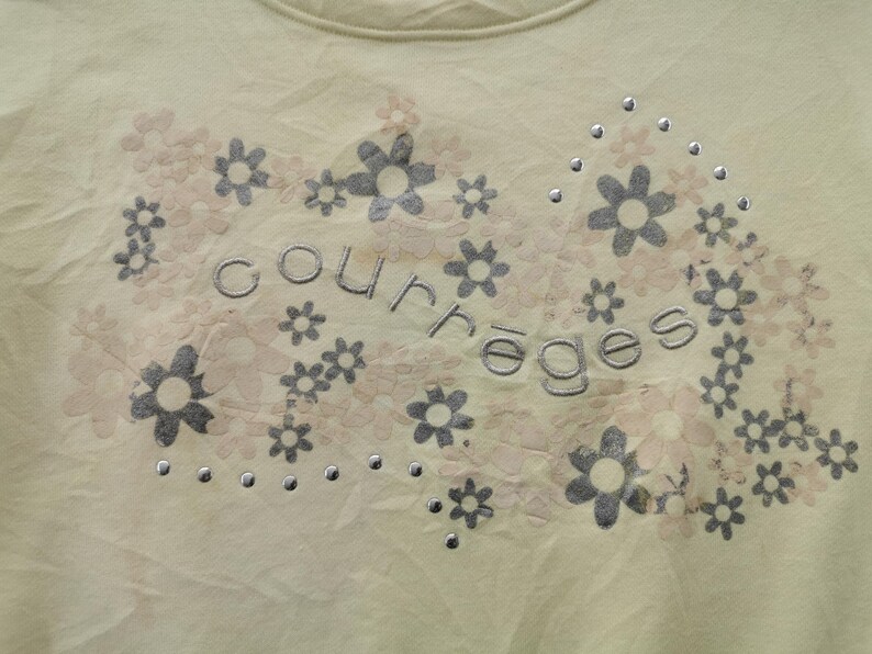 May include: A light yellow t-shirt with a floral design. The design includes gray and light pink flowers, with the word "courreges" in gray lettering. Small silver studs are scattered throughout the design.