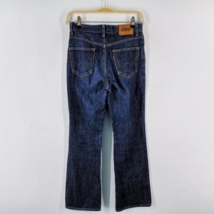 May include: Dark wash denim jeans with a flared leg, hanging on a wooden hanger. The back pockets feature a distinctive stitched design. A brown leather patch with the brand name "EDWIN" is visible at the waistband.