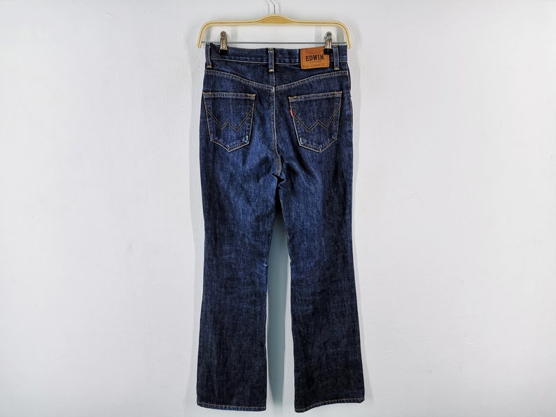 May include: Dark wash denim jeans with a flared leg, hanging on a wooden hanger. The back pockets feature a distinctive stitched design. A brown leather patch with the brand name "EDWIN" is visible at the waistband.