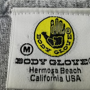 May include: Close-up of a gray fabric tag with the text "BODY GLOVE" in black. The tag features a yellow circle with a black hand logo and the text "BODY GLOVE". Below the logo is the text "Hermosa Beach California USA".