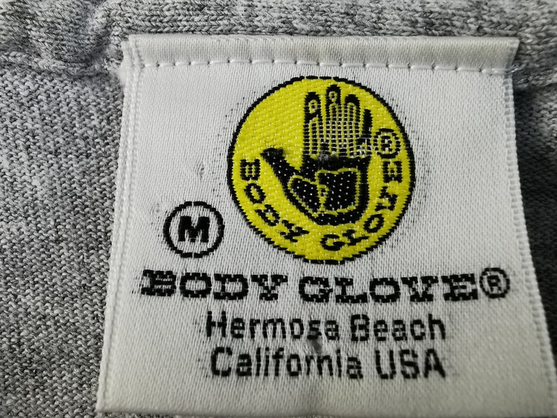 May include: Close-up of a gray fabric tag with the text "BODY GLOVE" in black. The tag features a yellow circle with a black hand logo and the text "BODY GLOVE". Below the logo is the text "Hermosa Beach California USA".