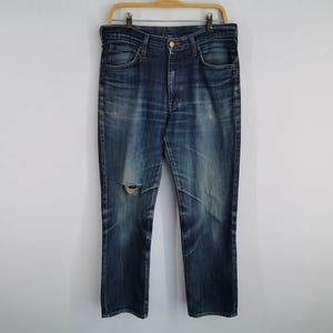K&ouml;nnte beinhalten: Dunkelblaue Jeans mit einem zerrissenen Knie-Detail, h&auml;ngen an einem Holzb&uuml;gel. Die Jeans haben ein klassisches F&uuml;nf-Taschen-Design und einen Knopf- und Rei&szlig;verschluss. Der Denim zeigt Ausbleichen und Abnutzung.