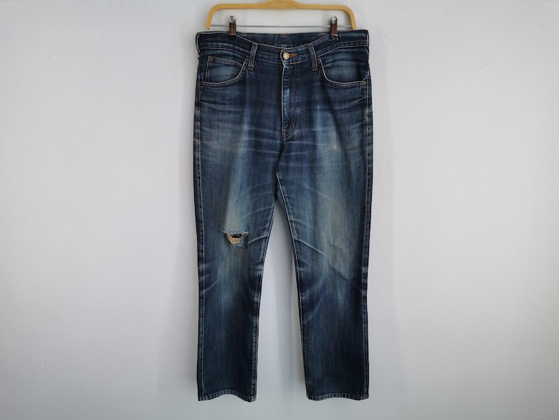 K&ouml;nnte beinhalten: Dunkelblaue Jeans mit einem zerrissenen Knie-Detail, h&auml;ngen an einem Holzb&uuml;gel. Die Jeans haben ein klassisches F&uuml;nf-Taschen-Design und einen Knopf- und Rei&szlig;verschluss. Der Denim zeigt Ausbleichen und Abnutzung.