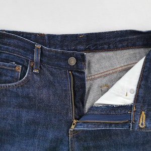 May include: Close-up of dark blue denim jeans with a partially unzipped fly. The jeans have a button closure, a small pocket on the front, and a classic five-pocket design. The interior lining is visible.