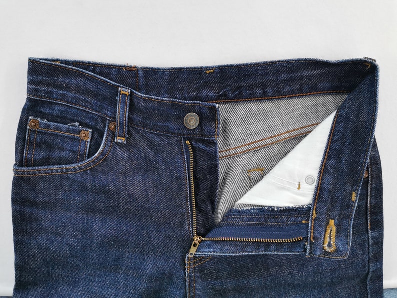 May include: Close-up of dark blue denim jeans with a partially unzipped fly. The jeans have a button closure, a small pocket on the front, and a classic five-pocket design. The interior lining is visible.