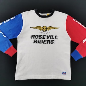 May include: White long-sleeve shirt with blue and red sleeves. The shirt features the text "ROSEVILL RIDERS" and a winged emblem. The sleeves have white text. The shirt has a black collar and red cuffs.