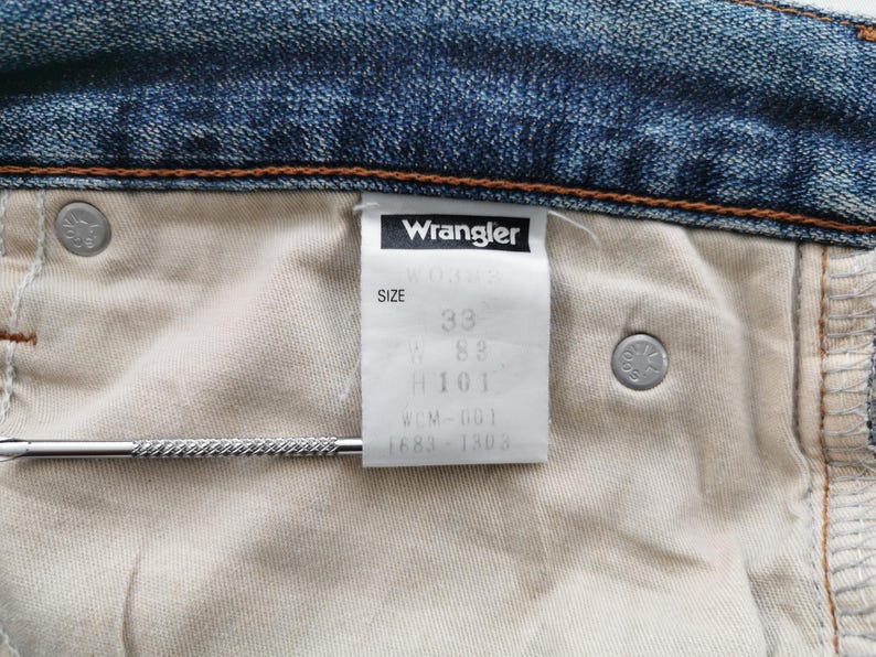 K&ouml;nnte beinhalten: Nahaufnahme der Innenseite einer blauen Jeans mit einem wei&szlig;en Etikett mit der Aufschrift "Wrangler" und der Gr&ouml;&szlig;e 33. Die Jeans hat einen Knopf und einen Metallstab.