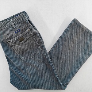 Op de afbeelding: Blauwe en grijze corduroy broek met verticale strepen. De broek heeft een kleine zak aan de achterkant en een kleine zak aan de voorkant. De broek is opgevouwen en plat op een wit oppervlak gelegd.