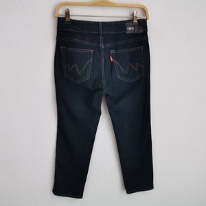 K&ouml;nnte beinhalten: Dunkelblaue Jeans aus Denim, die an einem Holzb&uuml;gel h&auml;ngen. Die Jeans haben ein klassisches F&uuml;nf-Taschen-Design mit einem roten Logo-Etikett und einem Lederaufn&auml;her mit dem Markennamen. Die Ges&auml;&szlig;taschen weisen ein markantes Nahtdesign auf.