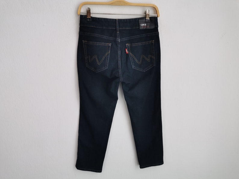 K&ouml;nnte beinhalten: Dunkelblaue Jeans aus Denim, die an einem Holzb&uuml;gel h&auml;ngen. Die Jeans haben ein klassisches F&uuml;nf-Taschen-Design mit einem roten Logo-Etikett und einem Lederaufn&auml;her mit dem Markennamen. Die Ges&auml;&szlig;taschen weisen ein markantes Nahtdesign auf.