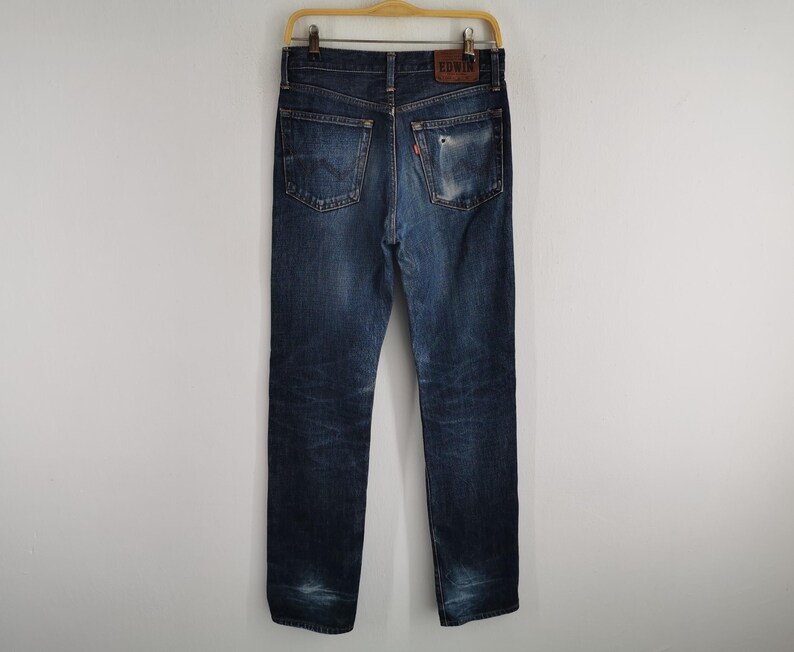 K&ouml;nnte beinhalten: Dunkelblaue Jeans mit Used-Look und klassischem F&uuml;nf-Taschen-Design. Die Jeans h&auml;ngen an einem Holzb&uuml;gel und zeigen die Ges&auml;&szlig;taschen und einen Leder-Markenaufn&auml;her.