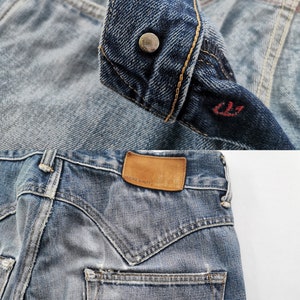 May include: Detailed shot of blue denim jeans, highlighting the button, pocket, and leather patch. The jeans have a faded look with visible stitching and a red tag. The leather patch reads "GENUINE QUALITY". The jeans are a classic style.