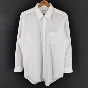 May include: A crisp white long-sleeve shirt with a classic collar and a chest pocket. The shirt is hanging on a wooden hanger against a dark background. The cuffs are buttoned, and the shirt appears to be made of a lightweight fabric.