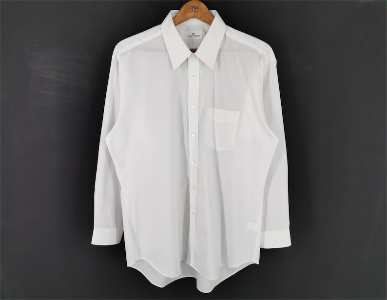 May include: A crisp white long-sleeve shirt with a classic collar and a chest pocket. The shirt is hanging on a wooden hanger against a dark background. The cuffs are buttoned, and the shirt appears to be made of a lightweight fabric.