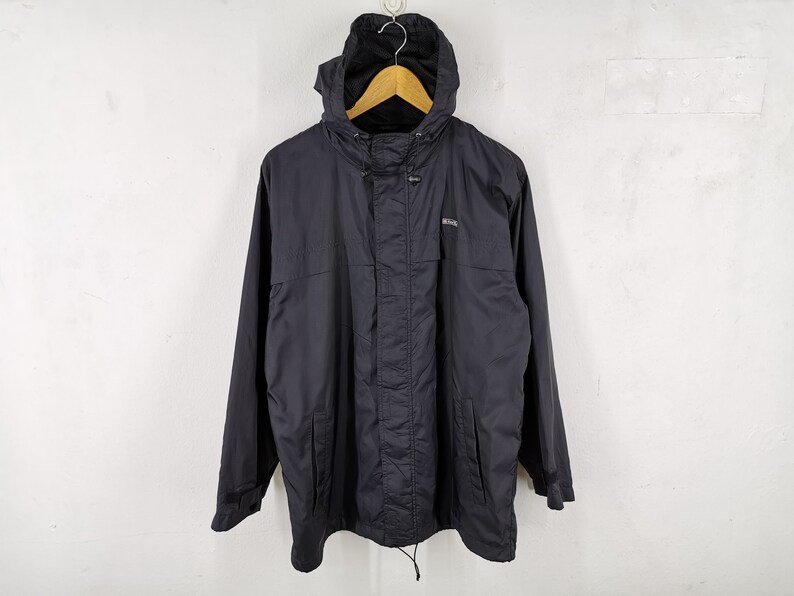 May include: A black hooded jacket with a drawstring at the waist and a small logo on the chest. The jacket is hanging on a wooden hanger against a white background. The jacket has a zipper closure and pockets.