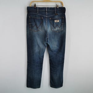 K&ouml;nnte beinhalten: Blaue Jeans aus Denim mit klassischem F&uuml;nf-Taschen-Design, h&auml;ngen an einem Holzkleiderb&uuml;gel. Die Jeans haben eine verblasste Waschung, einen Lederaufn&auml;her auf der Ges&auml;&szlig;tasche und das Wrangler-Logo. Die Jeans haben eine mittlere Waschung und scheinen einen geraden Bein-Stil zu haben.