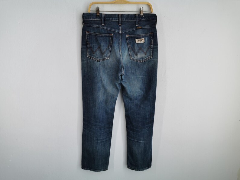 K&ouml;nnte beinhalten: Blaue Jeans aus Denim mit klassischem F&uuml;nf-Taschen-Design, h&auml;ngen an einem Holzkleiderb&uuml;gel. Die Jeans haben eine verblasste Waschung, einen Lederaufn&auml;her auf der Ges&auml;&szlig;tasche und das Wrangler-Logo. Die Jeans haben eine mittlere Waschung und scheinen einen geraden Bein-Stil zu haben.