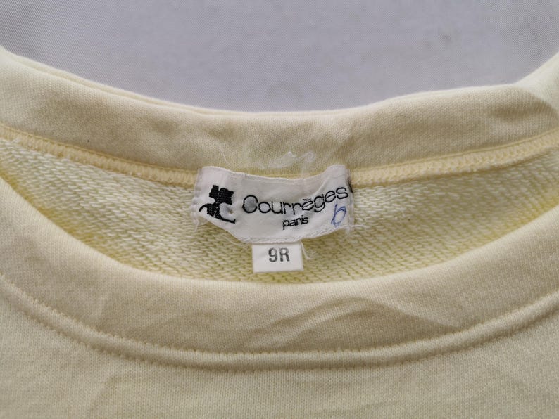 May include: Close-up of a pale yellow sweatshirt with a crew neck. A white label with the text "Courr&egrave;ges paris" and a logo is sewn into the neckline. The label also has the number "9R" printed on it.