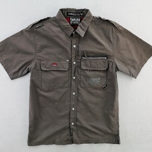 May include: A short-sleeved, button-down shirt in a muted gray-brown color. It features two chest pockets, one with a zipper, and various patches and printed text. The collar has a red and black plaid lining.