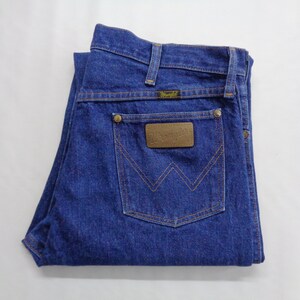 May include: Folded blue denim jeans with a brown leather patch on the back pocket. The patch has the brand name "Wrangler" embossed on it. The jeans have a classic five-pocket design and a button and zipper closure.