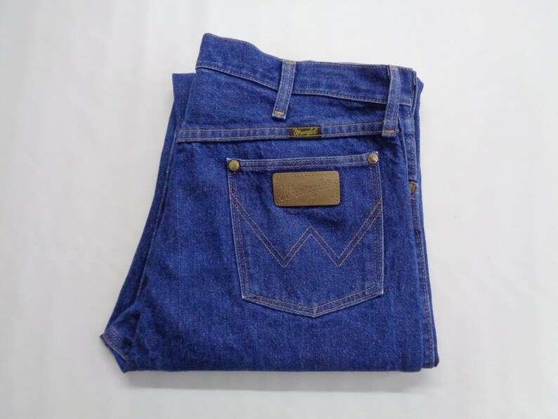 May include: Folded blue denim jeans with a brown leather patch on the back pocket. The patch has the brand name "Wrangler" embossed on it. The jeans have a classic five-pocket design and a button and zipper closure.