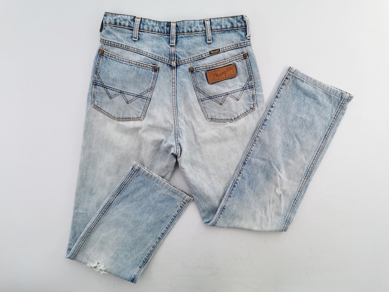 May include: Light blue denim jeans with a faded wash effect. The jeans feature classic five-pocket styling, a leather patch with the "Wrangler" logo, and a straight leg. The jeans have a distressed look with some wear and tear.