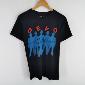 Op de afbeelding: Zwart T-shirt met het woord "DEVO" in rode letters boven een blauwe afbeelding van vijf figuren en een golvende lijn. Het T-shirt hangt aan een houten hanger tegen een witte achtergrond.