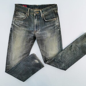 May include: Faded blue denim jeans with a classic five-pocket design. The jeans have a worn, distressed look with a small tear on the knee. The jeans are laid flat on a white surface.