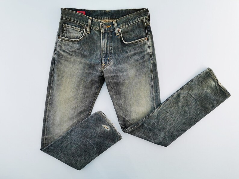 May include: Faded blue denim jeans with a classic five-pocket design. The jeans have a worn, distressed look with a small tear on the knee. The jeans are laid flat on a white surface.