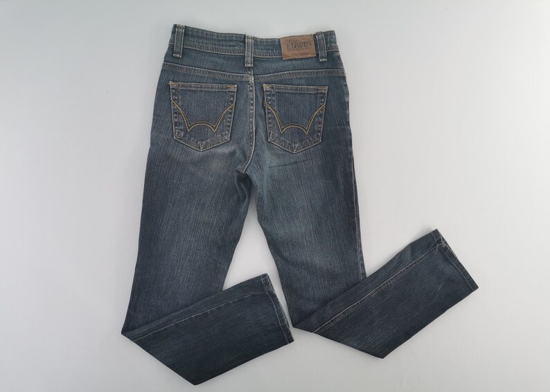 K&ouml;nnte beinhalten: Eine dunkelblaue Jeans aus Denim im klassischen F&uuml;nf-Taschen-Design mit einem braunen Lederaufn&auml;her am hinteren Bund mit der Aufschrift EDWIN und markanten goldfarbenen N&auml;hten auf den beiden Ges&auml;&szlig;taschen mit geschwungenem Bogenmuster.